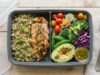 A meal prep box filled with fresh vegetables quinoa and grilled chicken on wood.